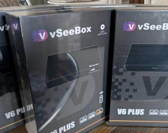 May include: Several black boxes of the VSeeBox V6 PLUS media player are stacked on a wooden surface. The boxes have the VSeeBox logo in purple and white, and the words "V6 PLUS" are printed in white.