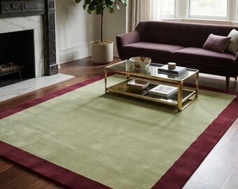 May include: A rectangular area rug featuring a large, solid light sage green center panel framed by a wide, deep burgundy border. The rug has a smooth, low-pile texture and is positioned flat on a dark wood floor in a living room setting.