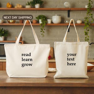 May include: Two off-white canvas tote bags with long handles. One bag has the words "read learn grow" printed in black. The other bag has the words "your text here" printed in black.