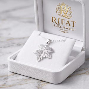May include: A silver leaf pendant on a delicate chain, displayed inside a white jewelry box. The interior lid features the gold-colored text RIFAT LUXE JEWELS EST. 2024 with a decorative emblem. The pendant has a textured, realistic leaf design.