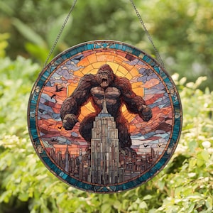 May include: Round stained glass window featuring a gorilla atop the Empire State Building, set against a sunset sky. The artwork includes a cityscape and birds, with a teal and brown border, suspended by a chain.
