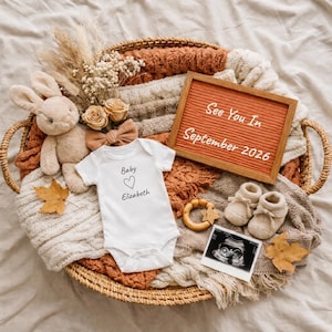 May include: A wicker basket filled with baby items, including a white onesie with "Baby Elizabeth" printed on it, a plush bunny, and a sign that reads "See You In September 2026". The arrangement includes baby booties, a wooden teether, and an ultrasound image.