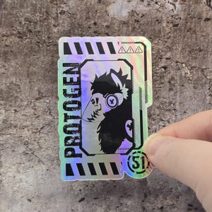 May include: A rectangular holographic sticker featuring a black silhouette of a creature with a visor and headphones. The text PROTOGEN is printed vertically on the left side, with the number 51 inside a circle at the bottom right corner.