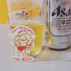Op de afbeelding: Een witte sticker met een cartoonkat die rode ondergoed draagt en de tekst "Don't mess with the best!" De sticker zit op een glas bier naast een blikje Asahi Super Dry bier.