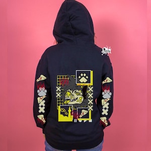 May include: A dark navy blue hooded sweatshirt featuring a graphic print on the back. The design includes a stylised animal character, pizza slices, paw prints, and yellow grid patterns. The sleeves also display repeating pizza slice graphics.