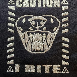 May include: A black patch with white stitching that reads "CAUTION I BITE" with a stylized snake head in the center.
