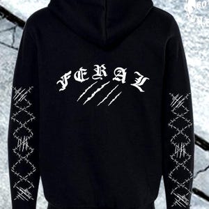 Puede incluir: Una sudadera con capucha negra que presenta el texto blanco FERAL en la parte superior de la espalda, sobre un gráfico de marcas de garras blancas. Ambas mangas están decoradas con un patrón blanco repetitivo de alambre de espino y garras.