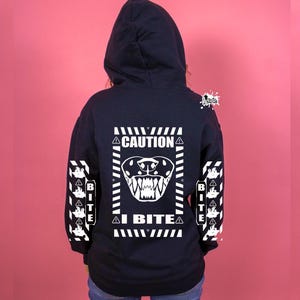 Puede incluir: Una sudadera con capucha negra con un gráfico blanco en la espalda. El gráfico es una señal de advertencia con una cara de animal gruñendo y el texto "I BITE". Las mangas tienen un patrón repetido de la señal de advertencia con el texto "BITE".