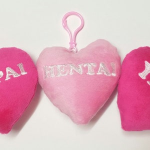 May include: Three pink heart-shaped plush keychains with silver embroidery. The keychains read "SENPAI", "HENTAI", and "YAOI".