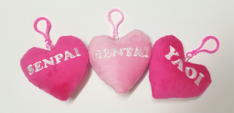 May include: Three pink heart-shaped plush keychains with silver embroidery. The keychains read "SENPAI", "HENTAI", and "YAOI".