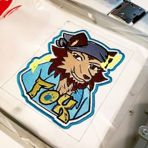 Puede incluir: Un parche bordado con un lobo de dibujos animados que lleva un pañuelo azul y una camisa azul con la palabra "Fox" en letras amarillas. El parche está sobre un fondo blanco.