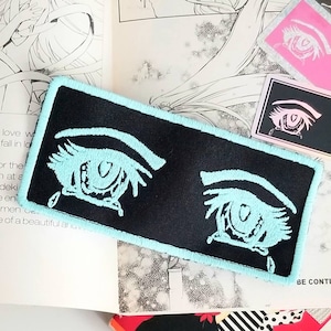 May include: A black fabric patch with two embroidered blue eyes with eyelashes and tears. The eyes have a heart shape in the centre of the iris.