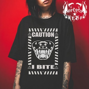 Puede incluir: Una camiseta negra con un estampado gráfico blanco de una boca de animal gruñendo con dientes afilados. El diseño está encerrado en un borde rectangular con rayas diagonales y el texto CAUTION en la parte superior e I BITE en la parte inferior.