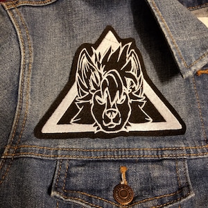 May include: A black and white embroidered patch of a wolf's head inside a white triangle on a blue denim jacket.
