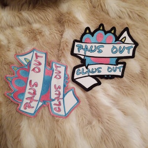 May include: Two embroidered patches with a paw print design. The first patch is pink, blue, and white with the text "PAWS OUT CLAWS OUT". The second patch is black, white, and pink with the text "PAWS OUT CLAWS OUT".