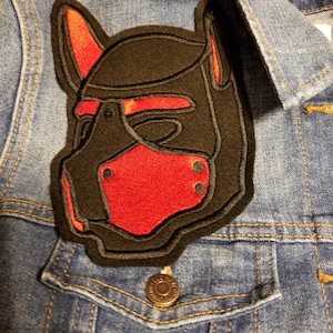 May include: Black and red embroidered patch of a dog muzzle. The muzzle is made of black fabric with red stitching and has a red nose and black eyes.