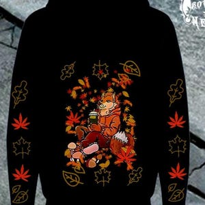 Peut inclure: Un sweat-shirt à capuche noir avec un motif central représentant un renard anthropomorphe portant un sweat orange et tenant un gobelet vert, entouré de feuilles d'automne rouges et orange. Les manches présentent un motif vertical de feuilles.