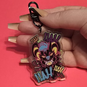 May include: A clear acrylic keychain with a cartoon monster design. The monster is black with yellow horns, blue and purple hair, and a menacing expression. The word "MAH" is written in blue and yellow. It is attached to a black metal clasp.