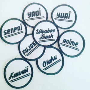 Puede incluir: Ocho parches redondos blancos bordados con texto negro. Los parches presentan las palabras "Senpai", "Anime", "Kawaii", "Fujoshi", "Weeaboo Trash", "Yai", "Yuri" y "Otaku".