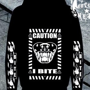 Puede incluir: Una sudadera con capucha negra que presenta un gráfico blanco en la parte posterior. El diseño incluye una cabeza de serpiente con colmillos expuestos dentro de un marco rectangular con el texto CAUTION e I BITE. Las mangas tienen el texto BITE.