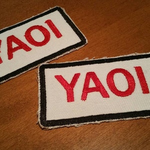 Puede incluir: Dos parches de tela blanca con bordes negros y texto bordado en rojo que dice "YAOI".