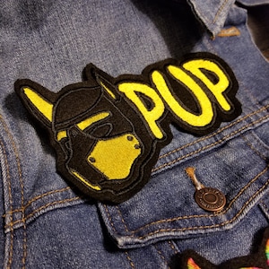 May include: A black and yellow embroidered patch with the word "PUP" in yellow lettering. The patch features a black dog mask with a yellow muzzle.