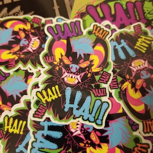 May include: A stack of colorful stickers featuring a cartoon hyena with a toothy grin and the word "HA!" in a graffiti style font. The stickers are on a green, black, and yellow background.