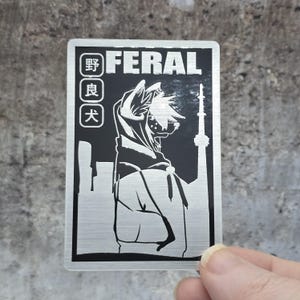 May include: A rectangular metallic sticker featuring a black and silver design. It displays the word FERAL in bold letters, three Japanese characters in vertical boxes, and a stylized illustration of a hooded figure standing before a city skyline.