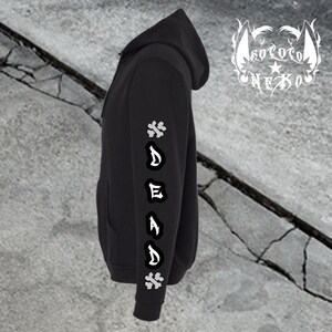 Puede incluir: Una sudadera con capucha negra que presenta un gr&aacute;fico vertical en la manga con la palabra DEAD en letras g&oacute;ticas blancas, flanqueada por dos s&iacute;mbolos de huesos cruzados blancos. Un logotipo blanco con el texto KOKORO NEKO es visible arriba a la derecha.