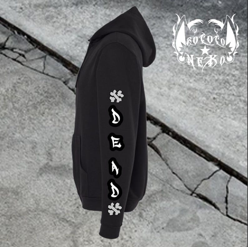 Puede incluir: Una sudadera con capucha negra que presenta un gr&aacute;fico vertical en la manga con la palabra DEAD en letras g&oacute;ticas blancas, flanqueada por dos s&iacute;mbolos de huesos cruzados blancos. Un logotipo blanco con el texto KOKORO NEKO es visible arriba a la derecha.