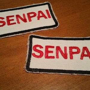Puede incluir: Dos parches de tela blanca con bordes negros y la palabra "SENPAI" bordada en rojo.