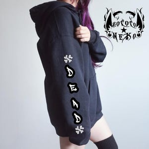 Puede incluir: Una sudadera negra con capucha que presenta un gr&aacute;fico blanco en la manga con la palabra DEAD escrita verticalmente en una fuente estilizada, flanqueada por dos s&iacute;mbolos en forma de cruz. Un logotipo con el texto KOTOTO NEKO y orejas de gato aparece.