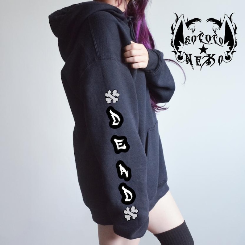 Puede incluir: Una sudadera negra con capucha que presenta un gr&aacute;fico blanco en la manga con la palabra DEAD escrita verticalmente en una fuente estilizada, flanqueada por dos s&iacute;mbolos en forma de cruz. Un logotipo con el texto KOTOTO NEKO y orejas de gato aparece.