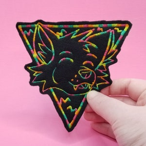 Peut inclure: Un patch brodé noir et blanc représentant le visage d'un animal de dessin animé avec une bordure triangulaire. La bordure est soulignée de vert néon, orange et rose.