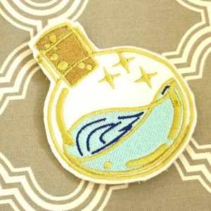 May include: An embroidered patch depicting a glass bottle with a cork stopper. The bottle contains a blue liquid with gold glitter inside. The bottle is outlined in gold thread.