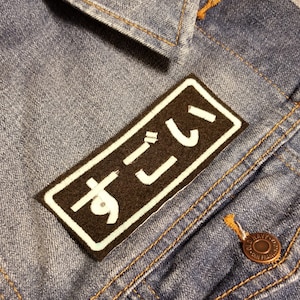 Puede incluir: Una chaqueta vaquera negra con un parche blanco que dice "すーごい" en japonés. El parche está cosido en el lado izquierdo de la chaqueta.