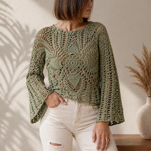 Könnte beinhalten: Ein salbeigrünes Häkeltop mit langen, ausgestellten Ärmeln. Das offene Design weist ein detailliertes Blumenmuster auf der Vorderseite auf. Das Oberteil wird mit hellen Hosen kombiniert. Der Stil ist böhmisch und handgefertigt.