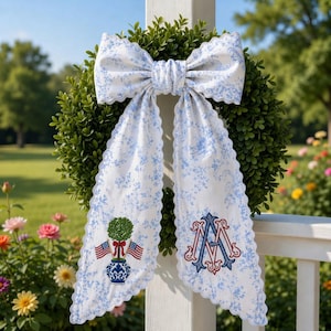 May include: A decorative wreath with a large bow made of white fabric with a blue floral pattern. The bow has a scalloped edge and is draped over a green wreath. The fabric streamers feature a topiary, American flags, and an embroidered monogram.