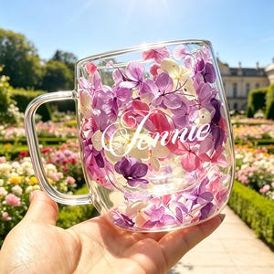 Könnte beinhalten: Ein transparenter doppelwandiger Glasbecher mit gepressten violetten, rosafarbenen und weißen Hortensienblütenblättern zwischen den Glasschichten und dem Namen Jennie in weißer Schreibschrift auf der Vorderseite des Bechers.