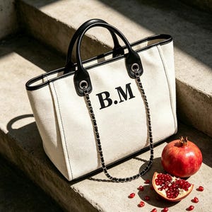 May include: A cream-coloured canvas tote bag with black leather trim and handles. The bag features the black monogram letters B.M on the front and includes a decorative black and silver chain strap. A whole and a sliced pomegranate are nearby.