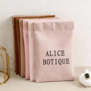 May include: A collection of soft, suede-like pouches in various shades of pink, brown, and tan. The front of the pink pouch features the text "ALICE BOTIQUE" in a dark font. The pouches are arranged vertically, with a gold-colored trim.