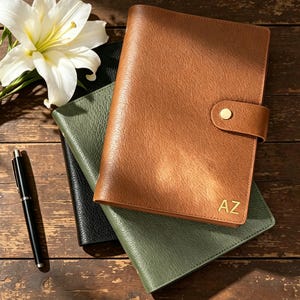 May include: A tan leather planner with a gold snap closure and the initials AZ embossed in gold on the bottom right corner. The planner is stacked on top of a green leather planner and a black leather planner, accompanied by a black pen.