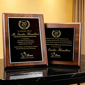 Op de afbeelding: Twee rechthoekige houten plaquettes met donkerbruine houtnerflijsten en zwarte middenpanelen. De plaquettes zijn voorzien van gouden tekst en een lauwerkransafbeelding met het getal 20, ter ere van Ms. Emilia Hamilton voor 20 jaar dienst bij het White Dove Medical Service Center.