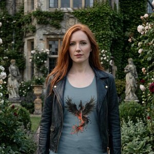 K&ouml;nnte beinhalten: Eine Frau tr&auml;gt eine schwarze Lederjacke und ein hellblaues T-Shirt mit einem schwarzen und roten Ph&ouml;nix-Design. Der Hintergrund zeigt ein Geb&auml;ude und einen Garten mit Statuen und Blumen.