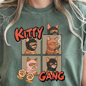May include: Sage green t-shirt featuring a graphic of four cats in balaclavas, with the words "Kitty Gang" in red and orange. The cats are depicted in various poses, some with weapons and one with a cigarette.