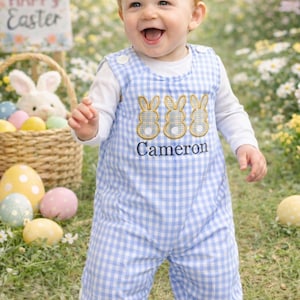 May include: A smiling toddler wearing a blue and white gingham romper with embroidered bunny designs and the name "Cameron". The child is in a grassy area with Easter decorations, including a basket, eggs, and a sign that says "Happy Easter".