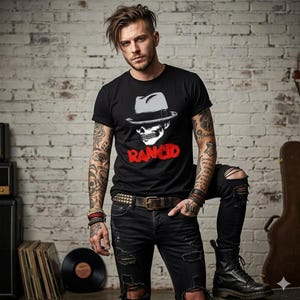 Puede incluir: Camiseta negra con una calavera con un sombrero fedora y la palabra "RANCID" en rojo. La persona lleva vaqueros negros rasgados, un cinturón con tachuelas y botas negras. El fondo es una pared de ladrillos.