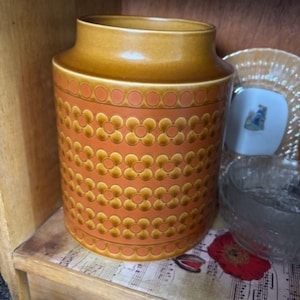 May include: A vintage ceramic jar with a warm orange base and a repeating floral pattern in shades of gold and brown. The jar has a rounded body and a slightly flared top, suggesting a storage container or decorative piece.