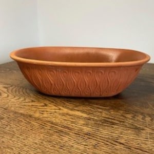 May include: A terracotta-colored, oval-shaped planter. The planter has a smooth interior and a decorative, repeating pattern of curved lines on the exterior. The rim is slightly raised, and the overall design is simple and elegant.