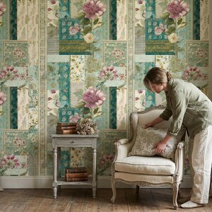 May include: A room with floral wallpaper in shades of teal, cream, and pink. A woman is arranging a decorative pillow on a cream-colored armchair. A small side table holds books and flowers. Beige curtains frame the scene.